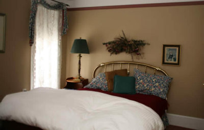 guest room with soft beige walls, brass bed , white, green and rust colored bed linens and window treatment