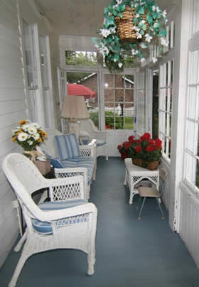 glass enclosed porch with white whicker couch and chair with blue stipped cussions facing the windows, flowering hanging basket hanging from the ceiling and red flowers on the white whicker coffee table